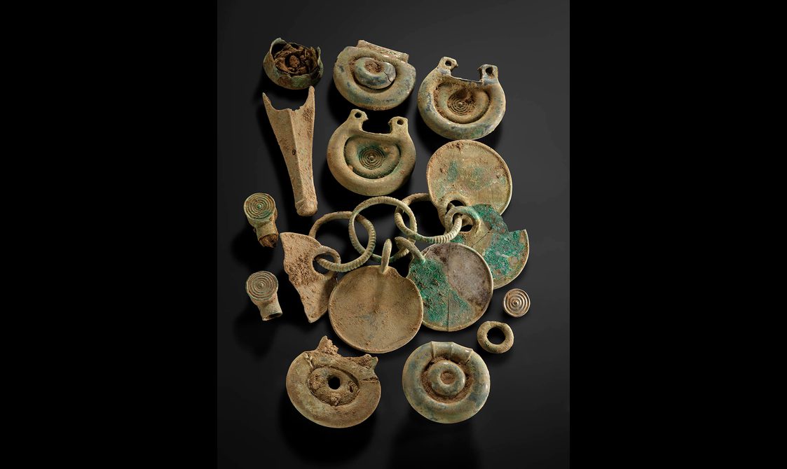 Twelve bronze objects, including some rounded fittings with concentric circle designs and a rattle pendant.