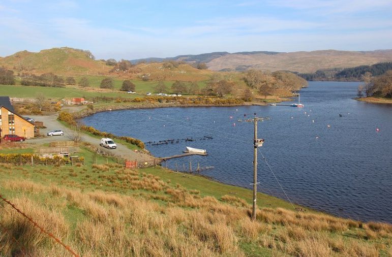 A lake surrounded by green and brown grassy hills with power lines and a house with cars visible to the left