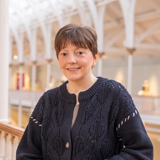 Angela Robb, a white cis woman in her early 40s, with short brown hair. Standing in front of railings in a light-filled museum atrium.