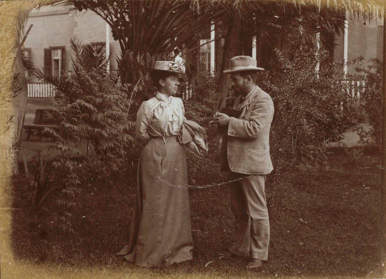 A black and white photograph of a man and woman talking to each other in a garden. The woman wearas a long skirt with a shirt and hat, the man wears a suit and hat.