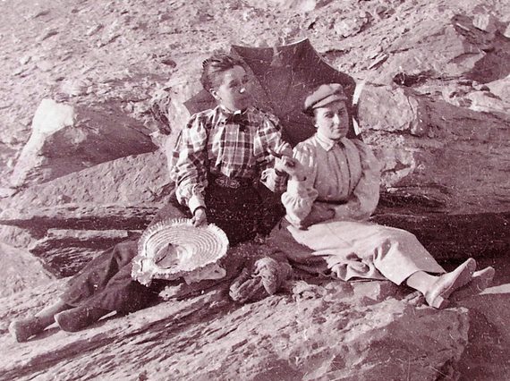 A black and white photograph of two women sitting on rocks in the desert. They are wearing buttoned up shirts and long skirts and are holding a parasol.