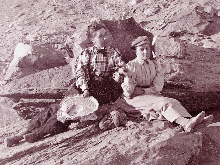 A black and white photograph of two women sitting on rocks in the desert. They are wearing buttoned up shirts and long skirts and are holding a parasol.