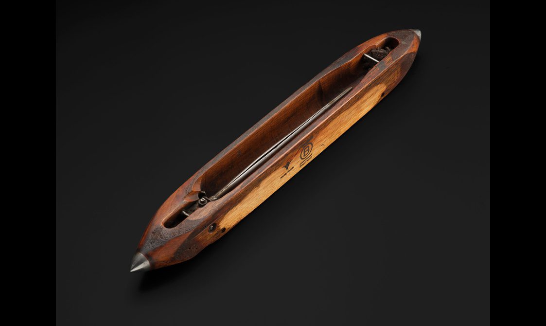 A wooden weaving shuttle. It is a small long object with a steal rod running through the middle.