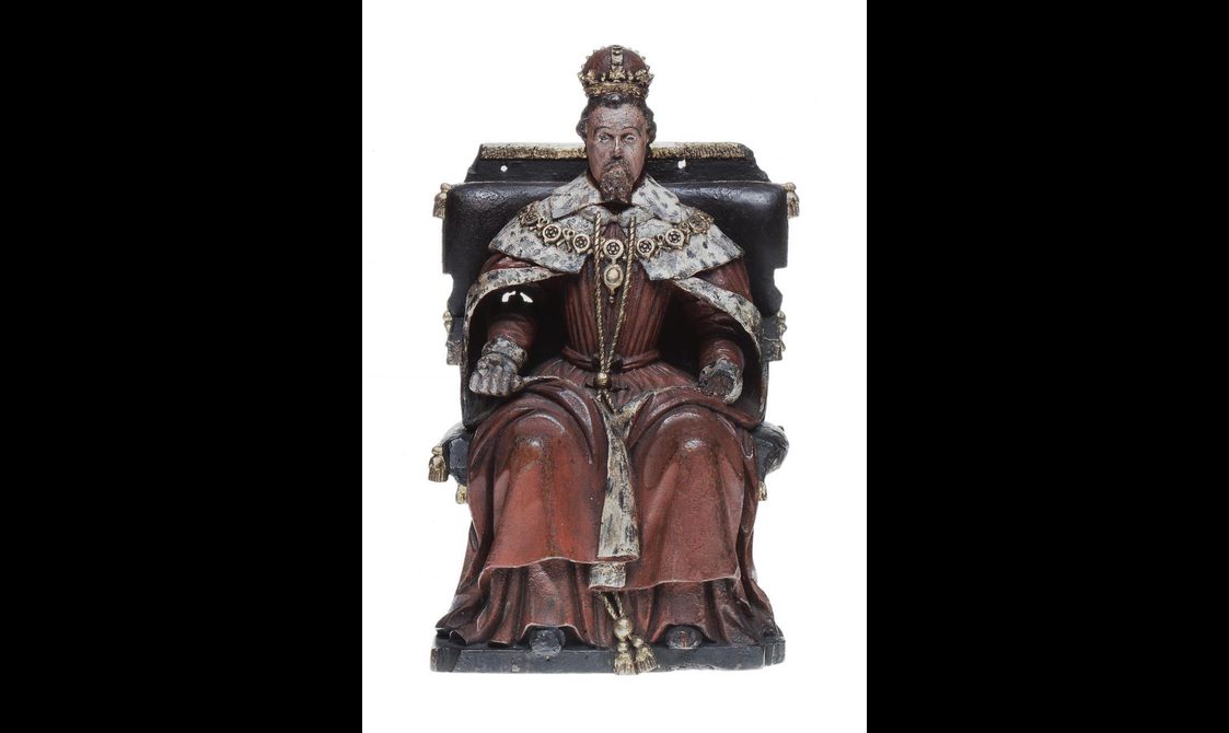 A wooden statue of a man sitting on a throne dressed as a king with rich red robes and a fur trimmed cloak wearing a crown.