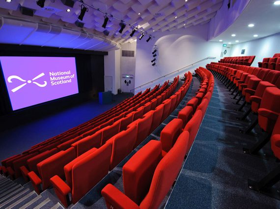 A large theatre space with raked seating and a big screen and lectern at the front. The seats are covered in red plush fabric and there is a National Museums Scotland logo on the screen.