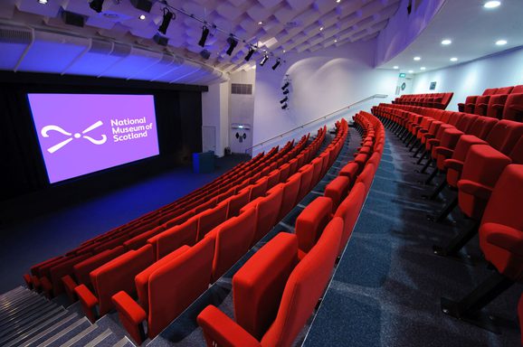 A large theatre space with raked seating and a big screen and lectern at the front. The seats are covered in red plush fabric and there is a National Museums Scotland logo on the screen.