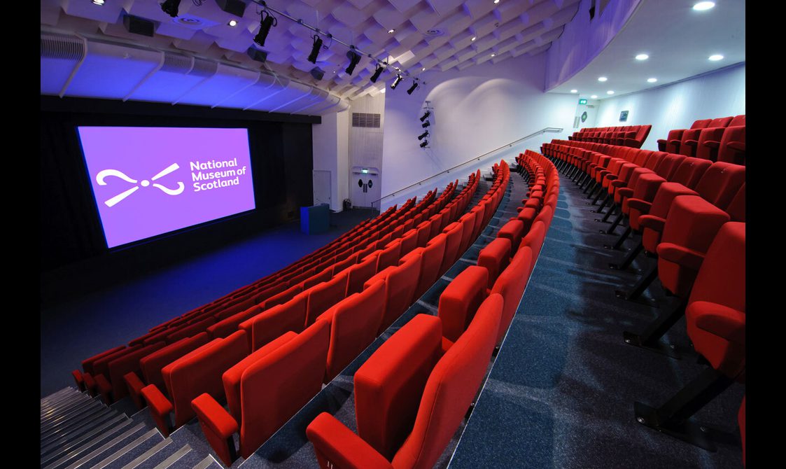 A large theatre space with raked seating and a big screen and lectern at the front. The seats are covered in red plush fabric and there is a National Museums Scotland logo on the screen.