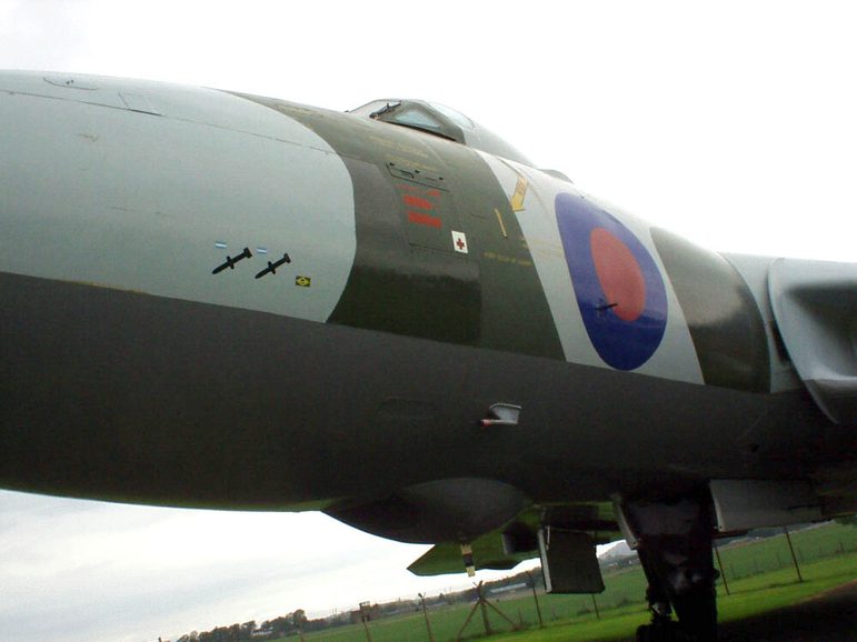 A close up of an aircraft that has a light grey and khaki green surface and two small dagger like marks on its nose.