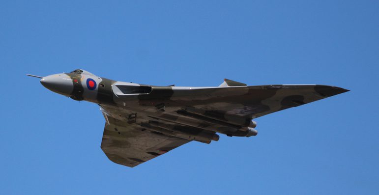 An aircraft with light grey and khaki green pattern on its surface flying in a blue sky.