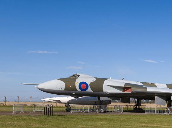 An aircraft with light grey and khaki green pattern over its surface and a blue and red round target near its nose. It sits on the ground surrounded by fencing in the countryside.