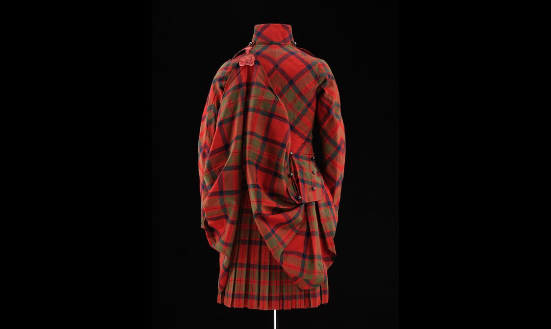 Back view of a mannequin wearing red tartan kilt with a matching jacket and pinned up tail of the jacket.