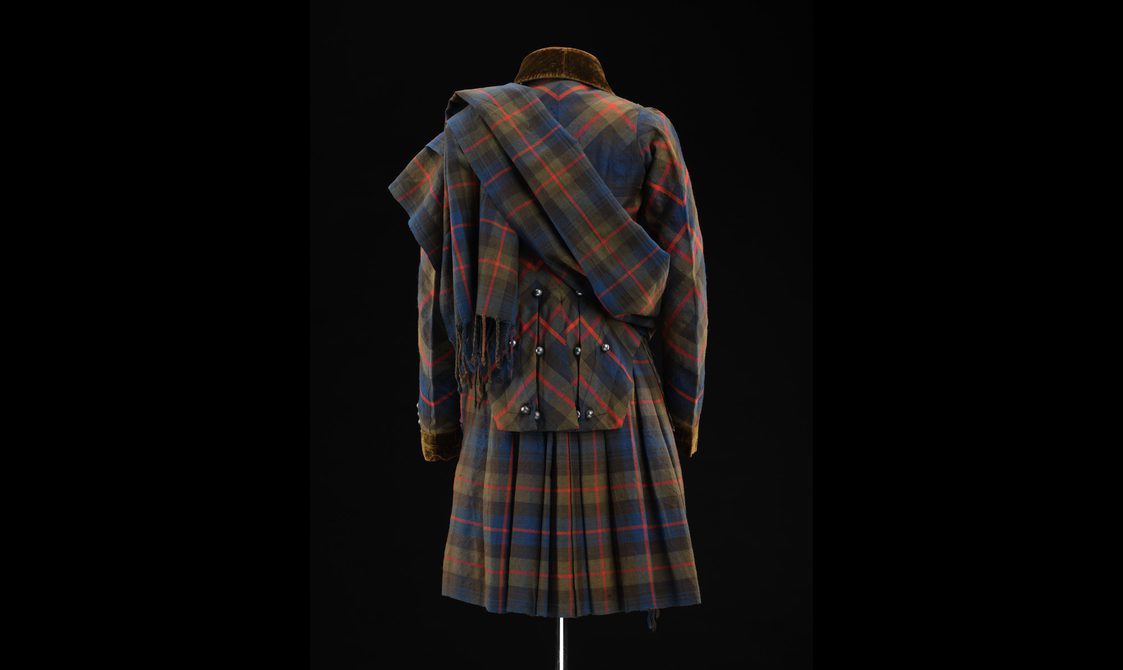 Back of a mannequin wearing a navy, green, and red tartan kilt with a matching jacket and draped shawl.