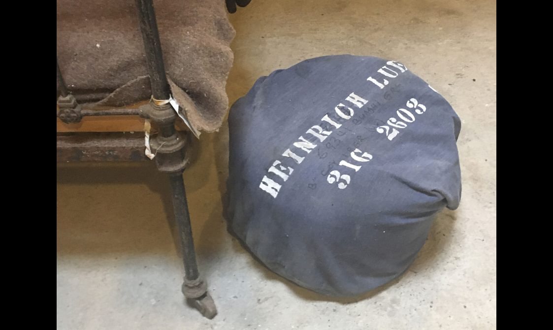 A blue bag at the side of a bed leg, with 'Heinrich Luckel 3I6G 2603' in white lettering.