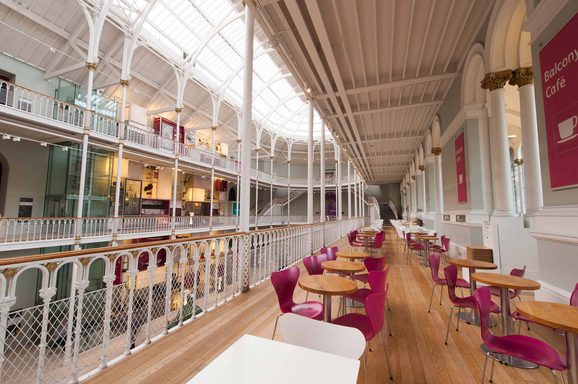 A series of cafe chairs and tables on a multi-level balcony looking out into a large atrium with a curved ceiling.