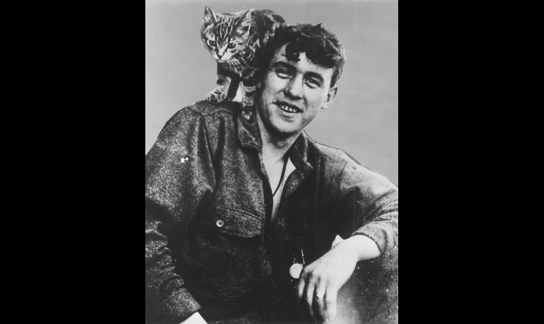 A black and white photograph of a man with short dark curly hair with a stripey cat on his shoulder.