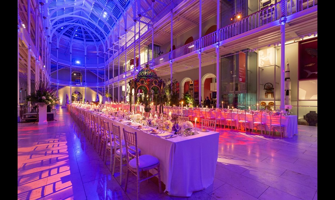 Long tables lined with chairs in a multi-level gallery space with balconies running around the outside. It is lit in pinks, blues, and purples.