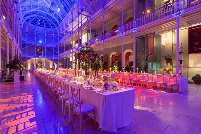 Long tables lined with chairs in a multi-level gallery space with balconies running around the outside. It is lit in pinks, blues, and purples.