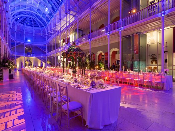 Long tables lined with chairs in a multi-level gallery space with balconies running around the outside. It is lit in pinks, blues, and purples.
