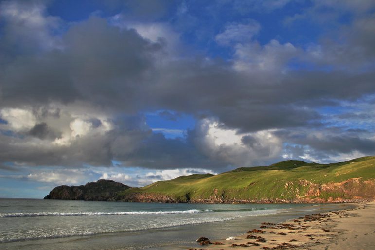 A beach with water lapping onto shore with green grassy cliffs in the background against a blue sky with grey clouds