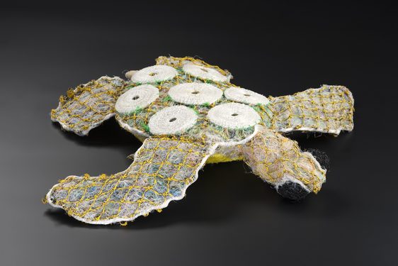 A turtle doll with a texture like a fuzzy teddy bear, made of compresses faded plastics contained within yellow netting. 7 white shell-like circles decorate its back.