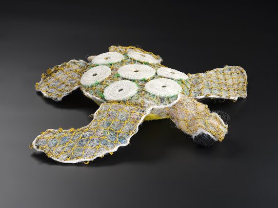 A turtle doll with a texture like a fuzzy teddy bear, made of compresses faded plastics contained within yellow netting. 7 white shell-like circles decorate its back.