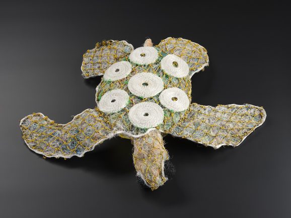 A turtle doll with a texture like a fuzzy teddy bear, made of compresses faded plastics contained within yellow netting. 7 white shell-like circles decorate its back.