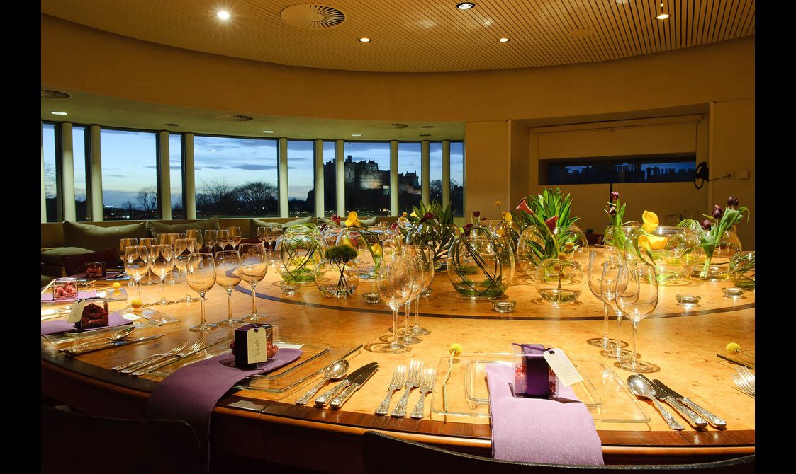 A large round room with windows overlooking Edinburgh Castle in the distance. There is a large round table set for dinner with decorative plants in the centre.