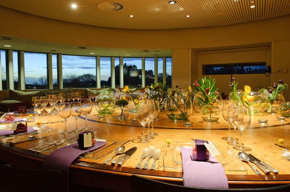 A large round room with windows overlooking Edinburgh Castle in the distance. There is a large round table set for dinner with decorative plants in the centre.