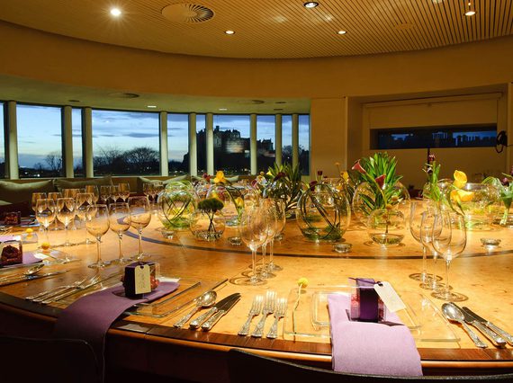 A large round room with windows overlooking Edinburgh Castle in the distance. There is a large round table set for dinner with decorative plants in the centre.