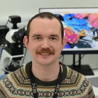 A head and shoulders photo of a man with short dark hair and a brown moustache, wearing a brown Fair Isle knit sweater and black work lanyard. He is sitting in front of a petrographic microscope, and a computer screen showing a microscope image of a rock thin section made up of blue, orange, and pink crystals.