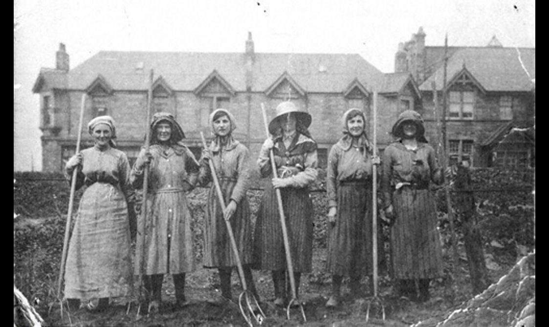 A black and white photograph of six women standing in a line holding rakes. They are in a field in front of a large building.