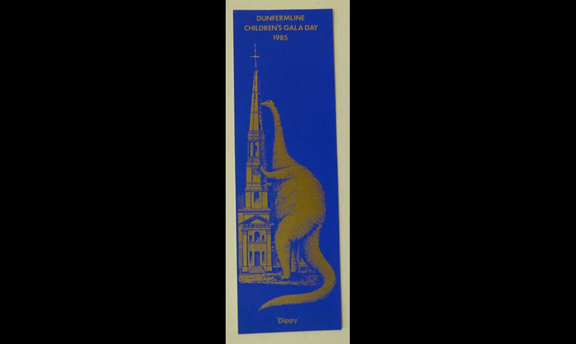A blue bookmark with gold design. 'Dunfermline children's gala day 1985' An image of 'Dippy' the dinosaur climbing Dunfermline Abbey.