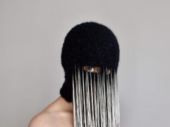 A person wears a black mask that covers their face except for their eyes. There are long white and gret threads attached from the eye openings.