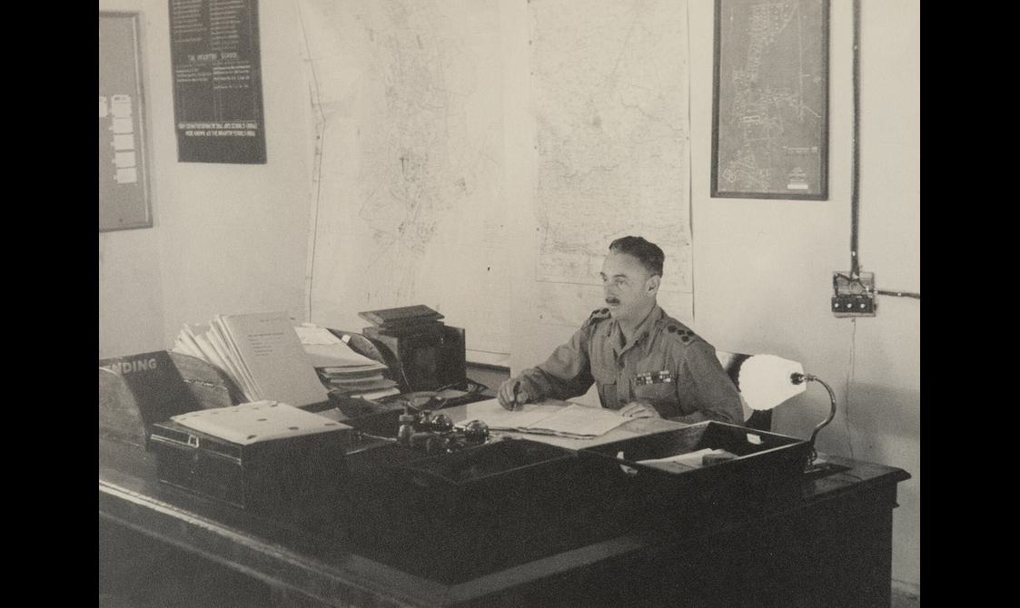 A man in army uniform sitting at desk stacked with books and papers
