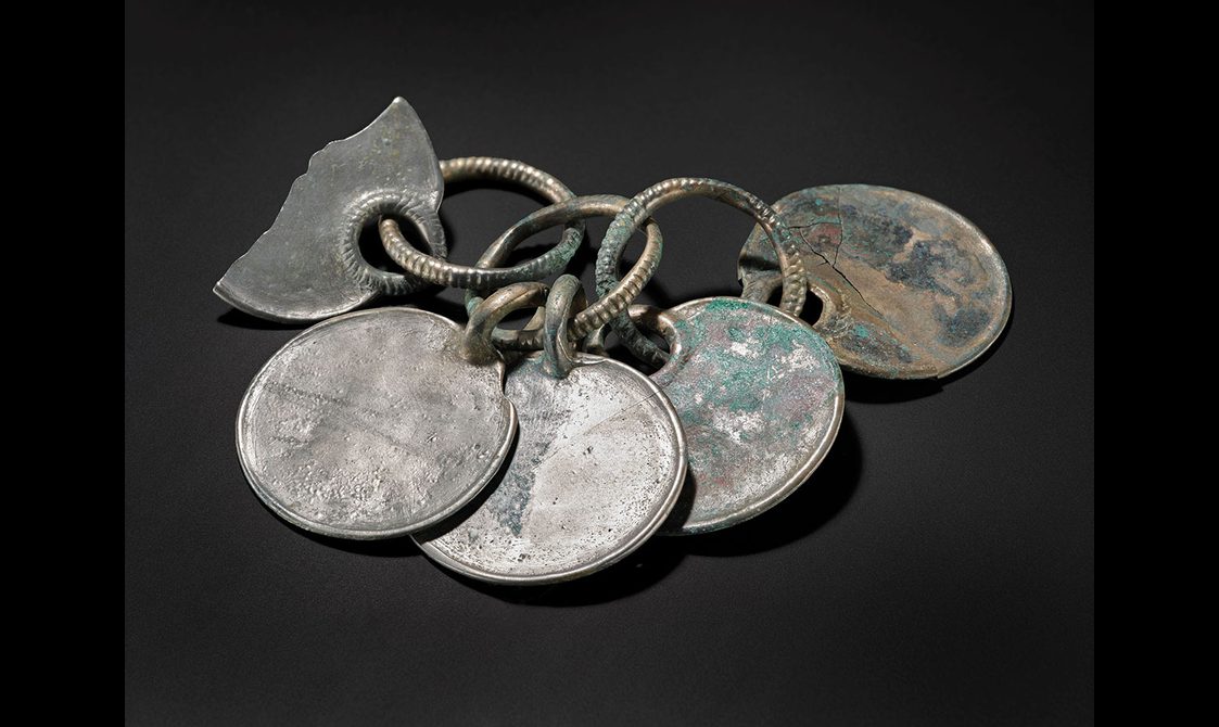 Three rings linked together in a row. Five bronze disks are hanging from these rings, creating a rattle. The surface is silvery with some green corrosion.