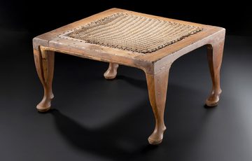 A wooden stool with a woven pad on top, photographed against a black background.