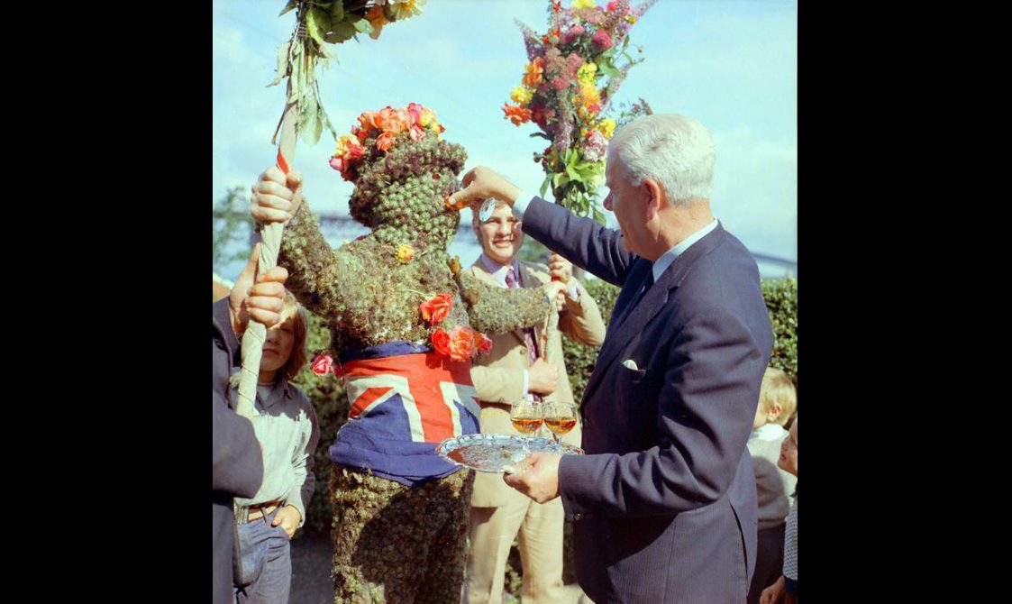 A figure in a costume made of plants with a floral hat on its head and a union jack flag wrapped around its waist, being fed a small glass of whisky by a man in a suit.