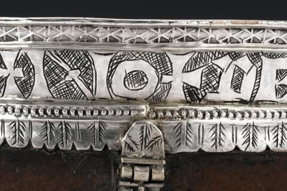 A wide strip of polished silver with a smooth top edge and a scalloped bottom edge. There are old letters engraved the full length of the piece bordered by engraved patterns.