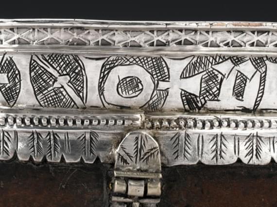 A wide strip of polished silver with a smooth top edge and a scalloped bottom edge. There are old letters engraved the full length of the piece bordered by engraved patterns.