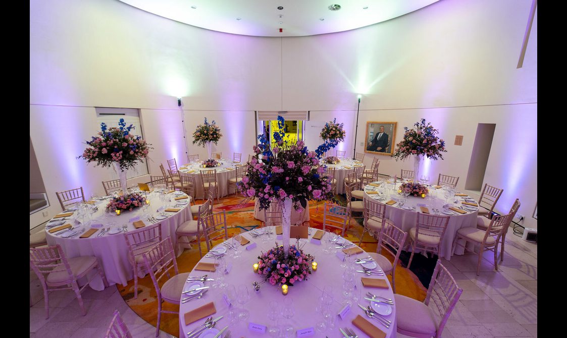 Tables set up for a meal in a circular room with white walls and purple lights.