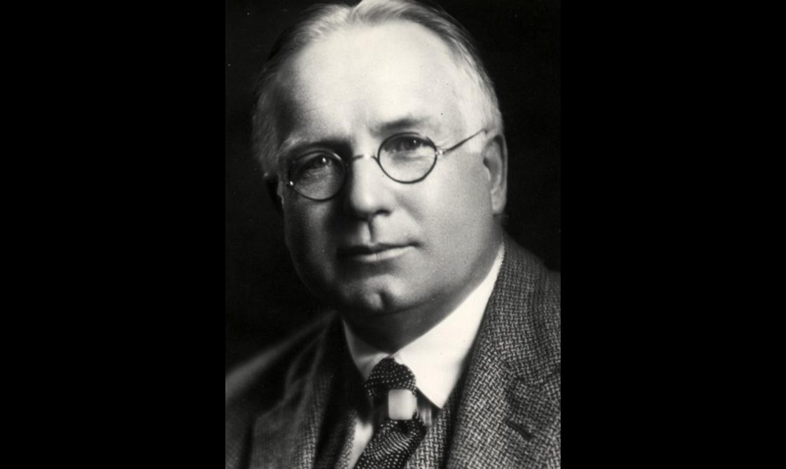 Black and white headshot of a man with thin white hair and round glasses.