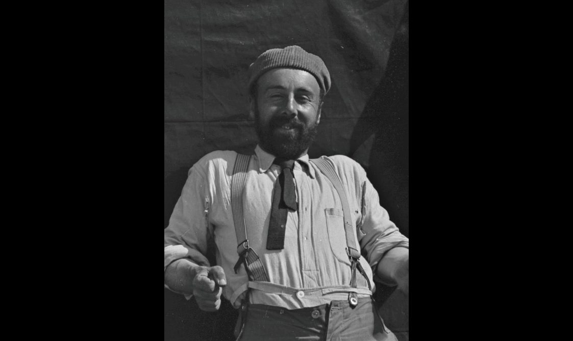 Black and white photo of a smiling man with a dark beard wearing a corduroy hat and suspenders.