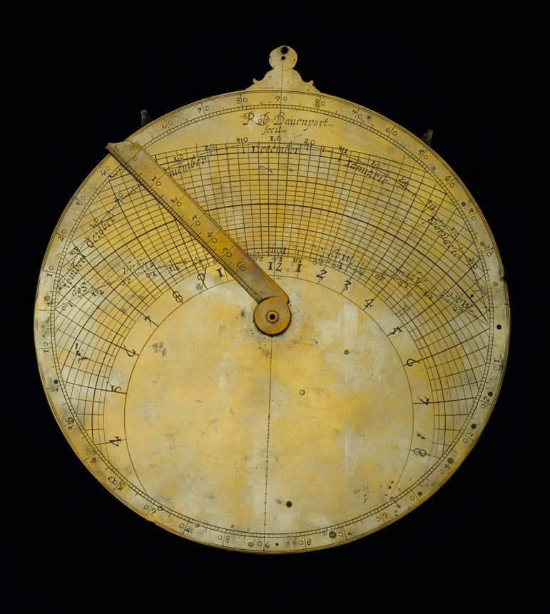 Round flat bronze metal disc with markings and a rotating arm attached at the centre.