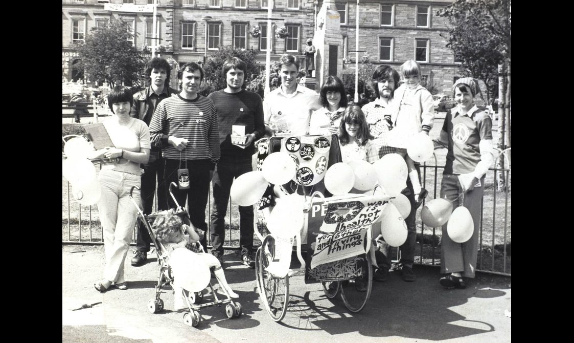 A black and white photo of a group of people standing in a city square holding balloons. There is a child's pram covered in protest signs and buttons.