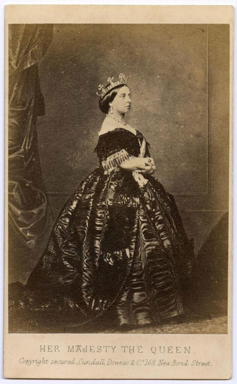 A black and white postcard of Queen Victoria in a dark dress with a fitted bodice and full skirt. She is wearing a crown and jewellery and is standing in profile.