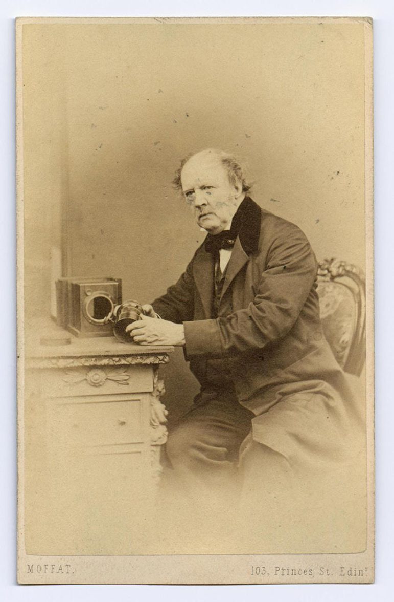 A faded black and white photograph of a man in a dark suit sitting on a chair at a desk holding the parts of an old camera
