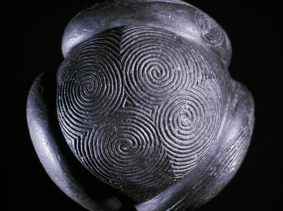 A close up of a grey ball with four circular disks attached to it, set against black background. Decorated with spiral carvings.