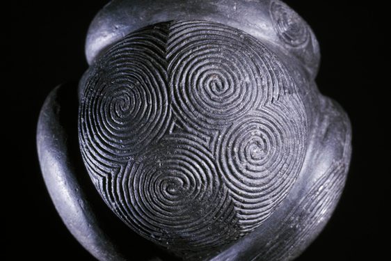 A close up of a grey ball with four circular disks attached to it, set against black background. Decorated with spiral carvings.