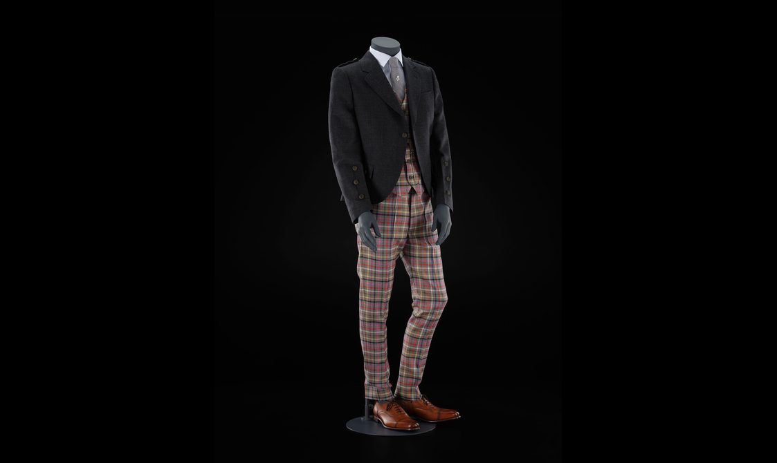 A mannequin wearing colourful tartan trousers, a dark wool suit jacket and tie with brown leather shoes.