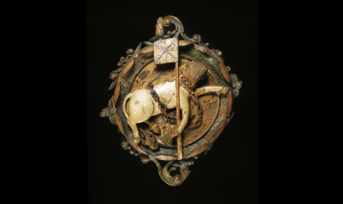 A round wooden sculpture of a unicorn folding a flag with a decorative boarder